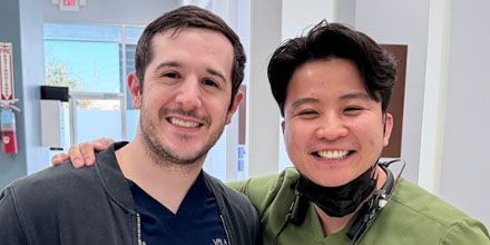 Two smiling people posing indoors, one in a dark shirt and one in a green shirt with headphones around the neck