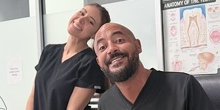 Two smiling people in black scrubs posing in a clinic hallway, with a dental anatomy poster behind them