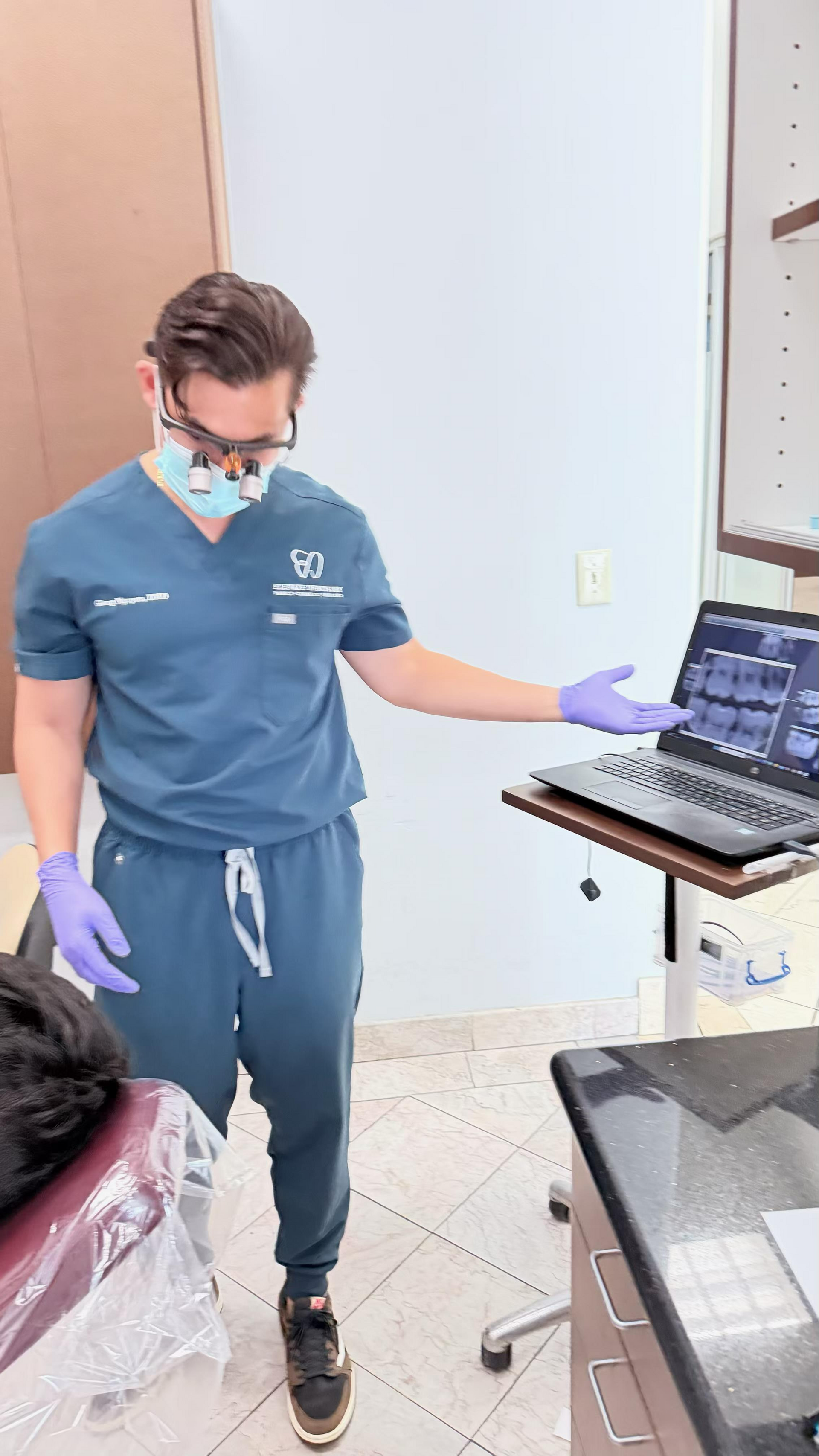 Medical professional in blue scrubs and gloves stands beside a laptop on a cart in a clinical room.
