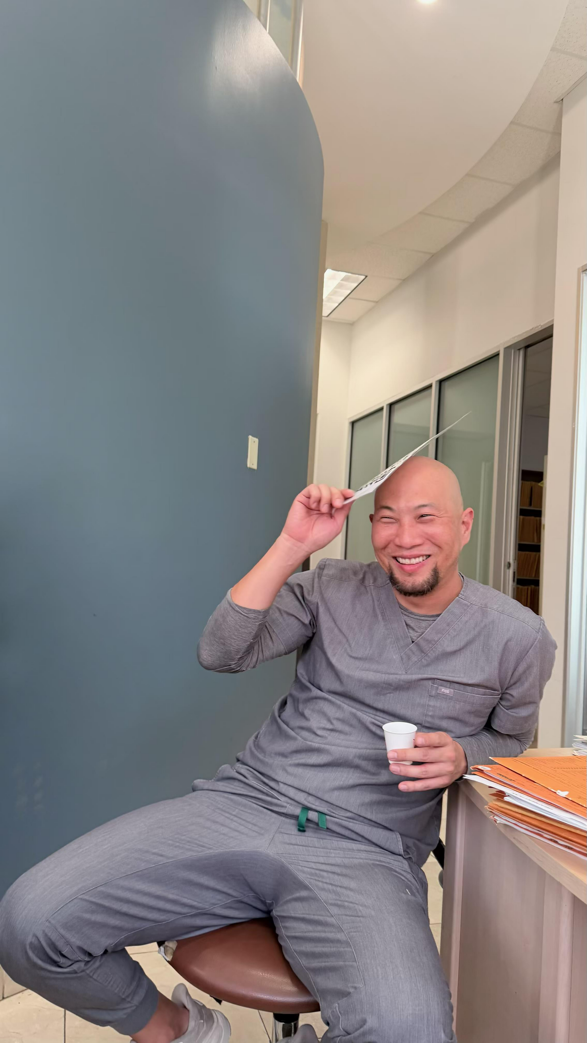 Smiling person in gray clothes sits by a blue wall, holding a cup and waving in a hallway.