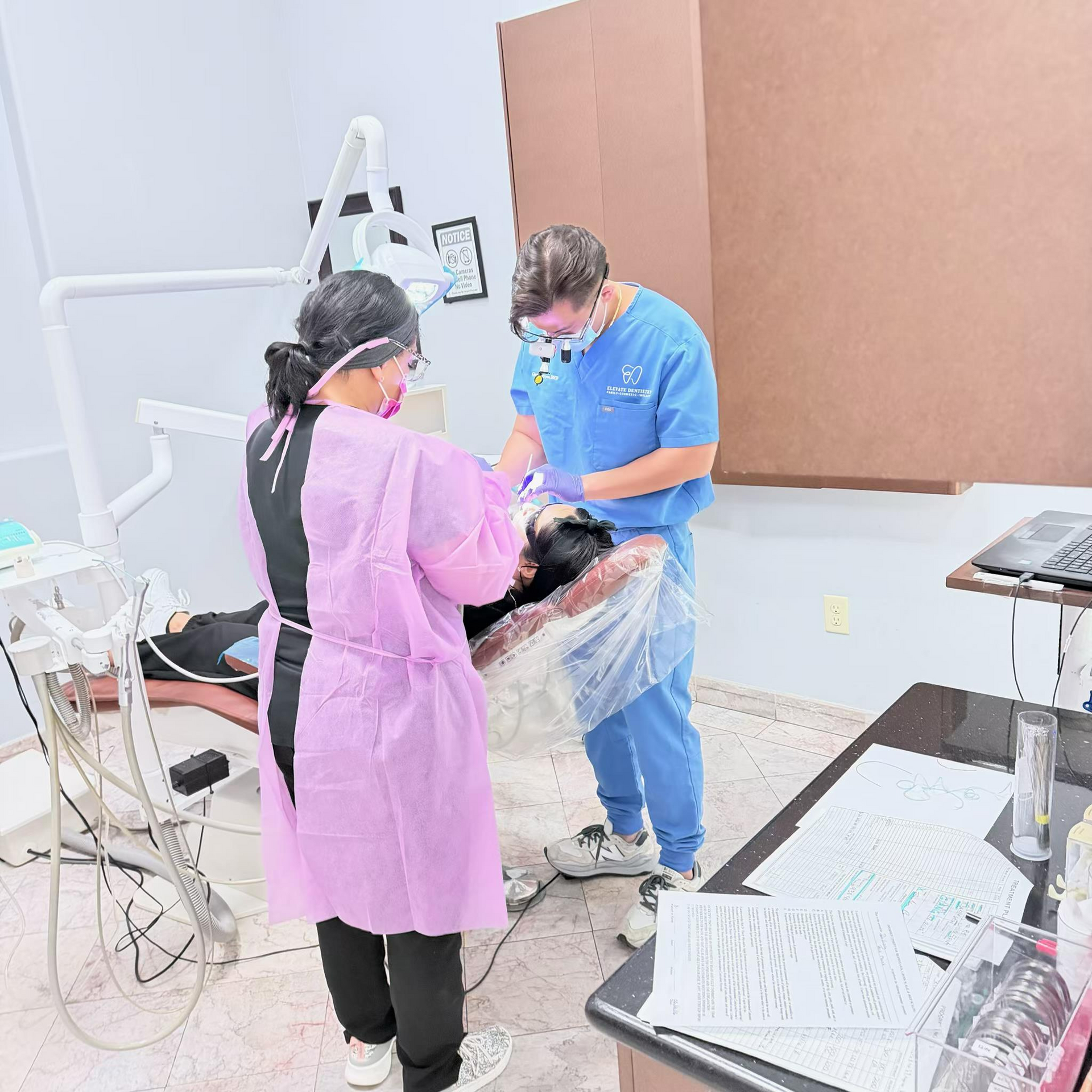 Dental staff treating a patient in a clinical exam room, with one person wearing blue scrubs and another in a pink coat.