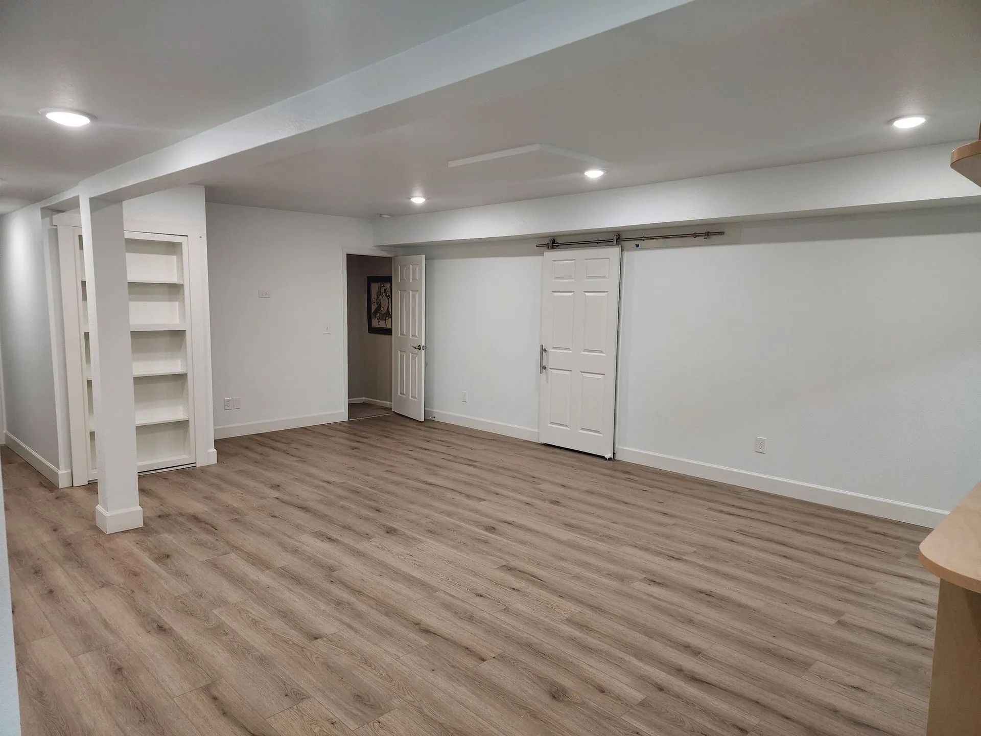 Empty basement room with light wood flooring, white walls, recessed lights, and built-in shelves.
