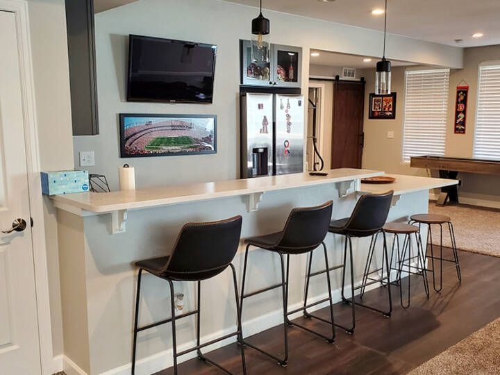 Modern kitchen bar with four black stools, white countertop, wall TVs, and pool table in the background