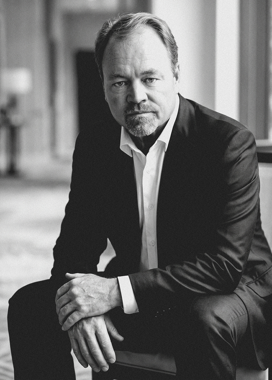 Black-and-white portrait of a man in a dark suit seated with hands folded, looking at the camera.