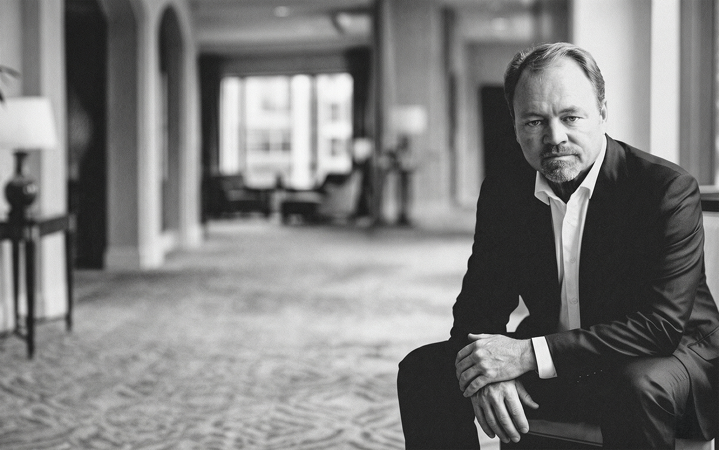 Black-and-white portrait of a man in a suit seated in a corridor, looking at the camera.
