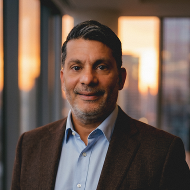 Portrait of a man in a brown blazer and blue shirt, standing indoors with a blurred sunset-lit background