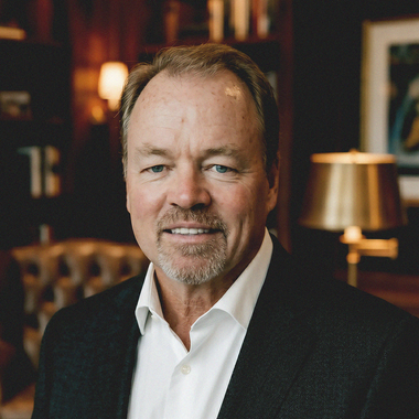 Smiling man in a dark blazer and white shirt in a warmly lit room with a lamp and bookshelves.