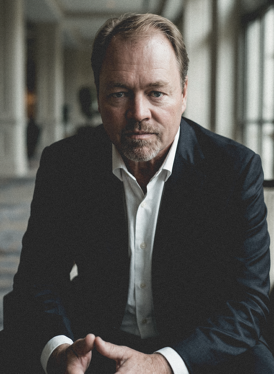 Man in a dark suit seated indoors, leaning forward with hands clasped, looking at the camera.