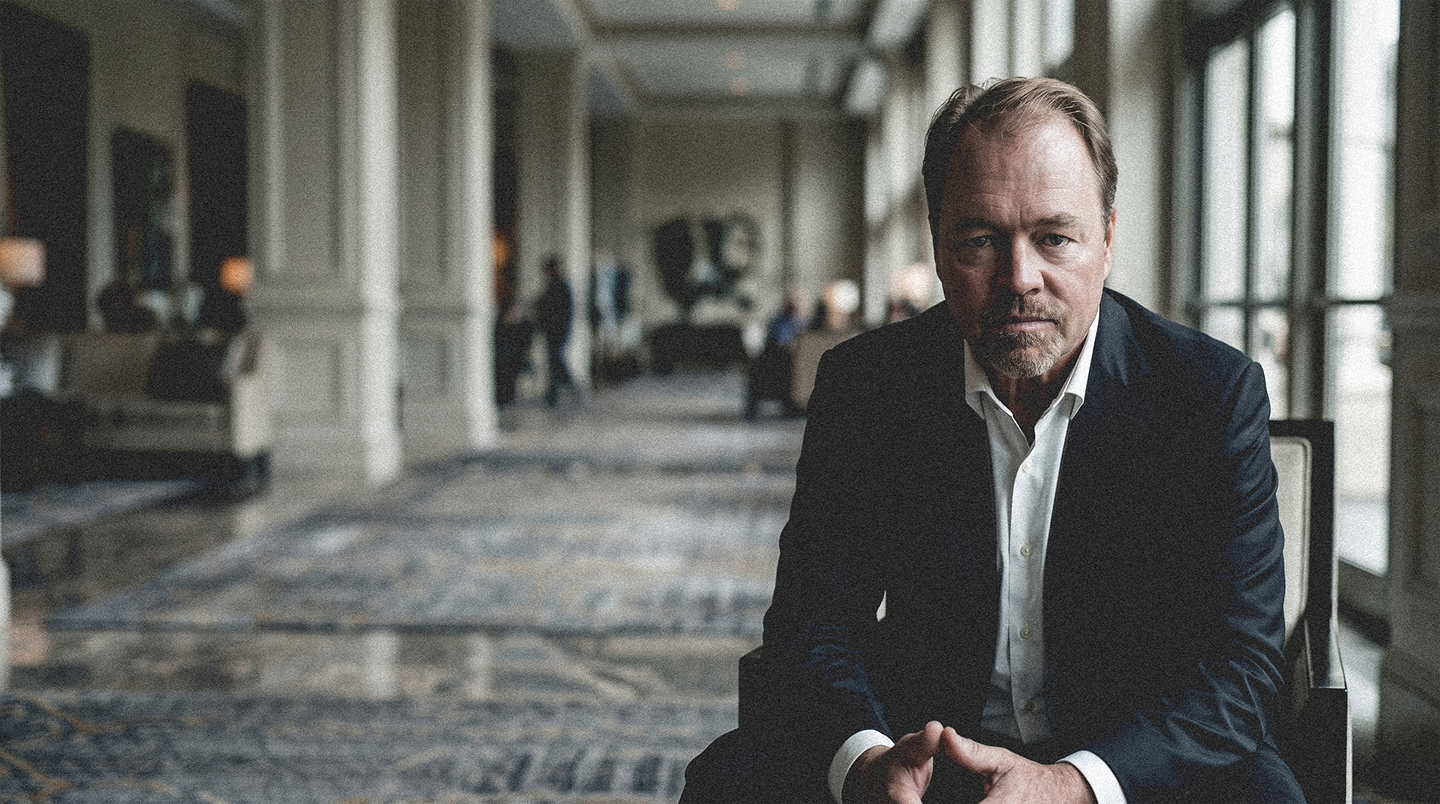 Man seated in a dim hotel lobby, wearing a dark suit and looking toward the camera.