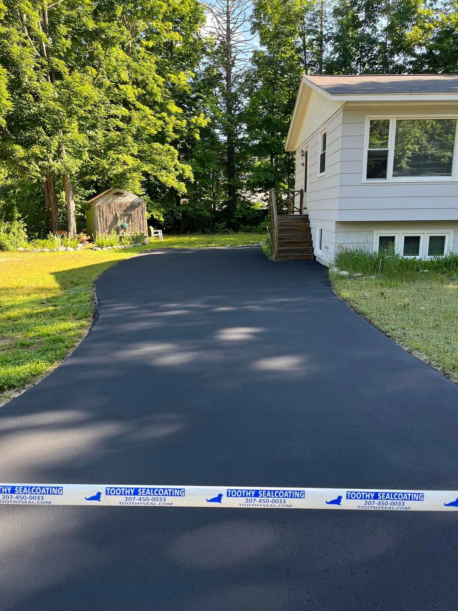 Freshly paved black driveway beside a white house and green yard, with caution tape in the foreground