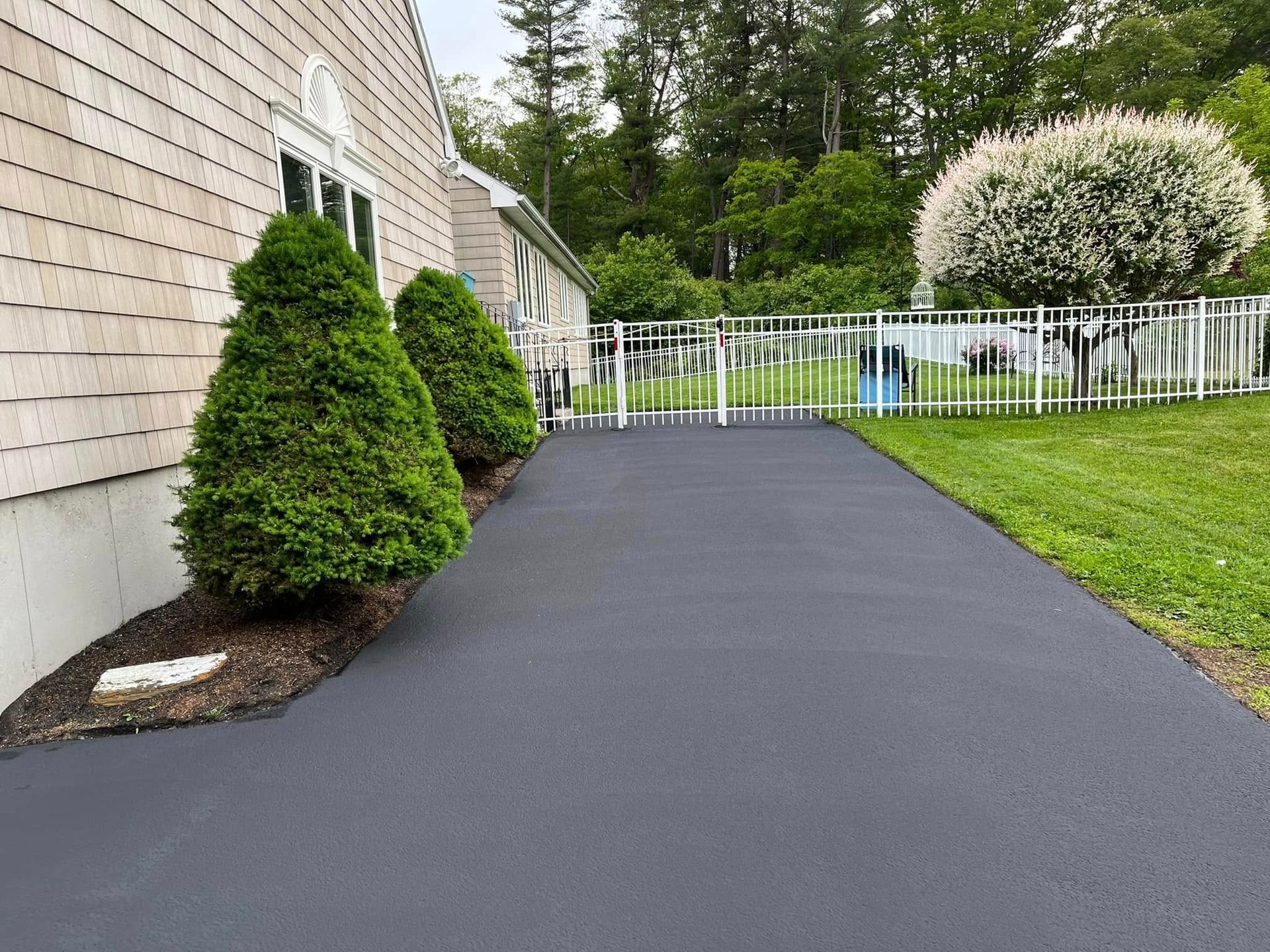 Driveway beside a house with trimmed shrubs and a white fence leading to a backyard.