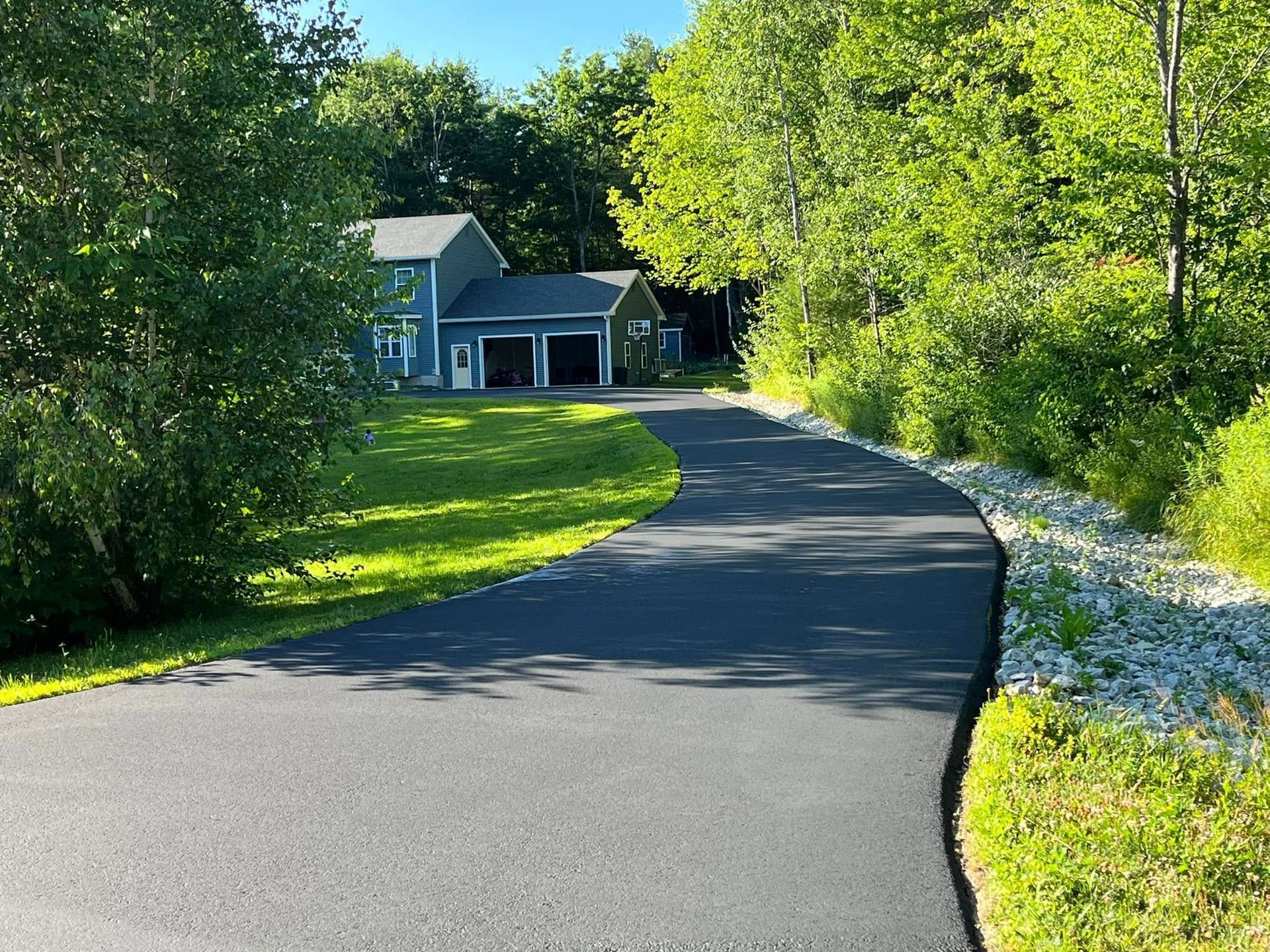 Curving driveway leading to a house, lined with lush green trees and grass.