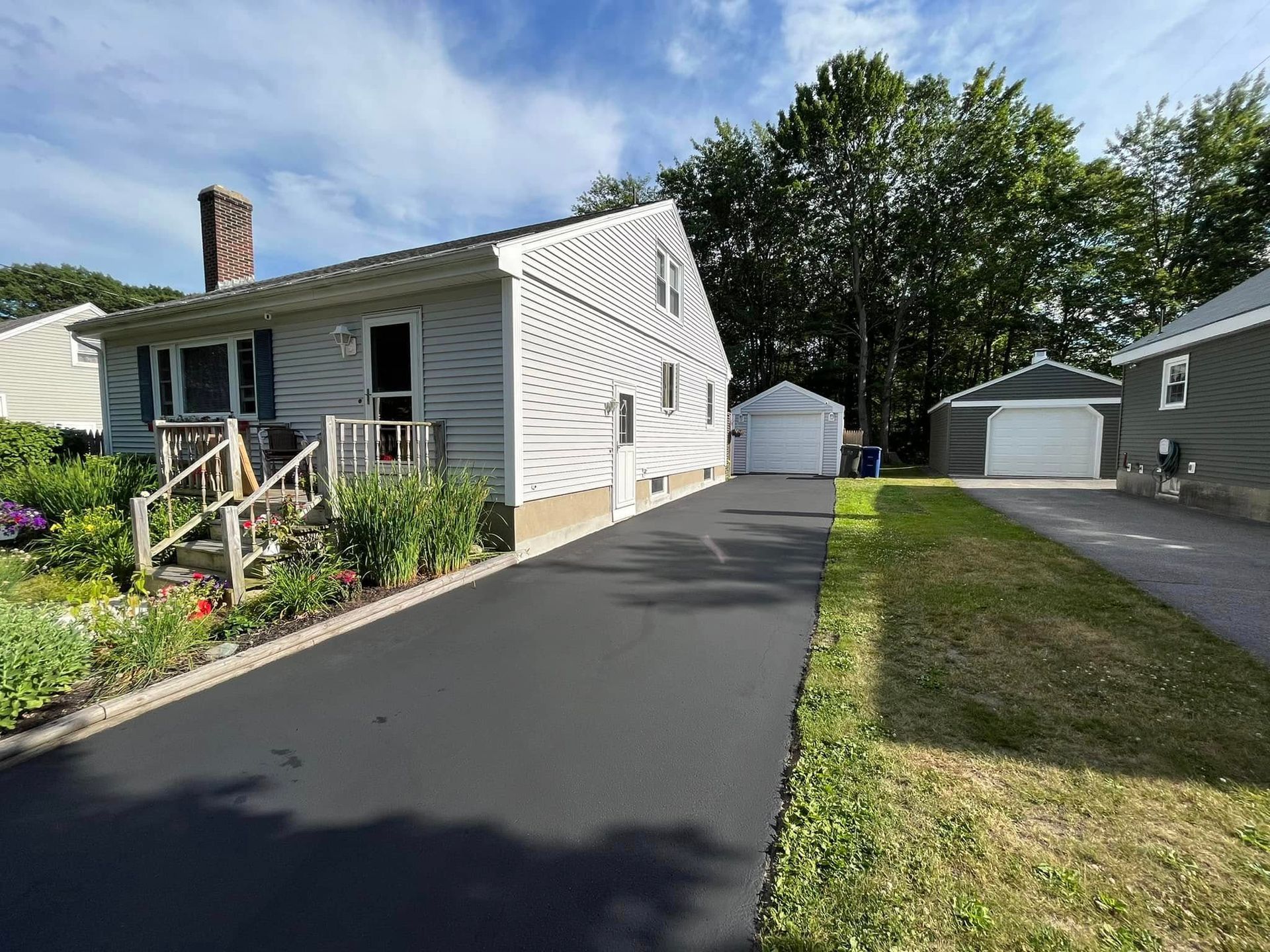 White house with a black driveway, green lawn, and detached garage on a sunny residential street.