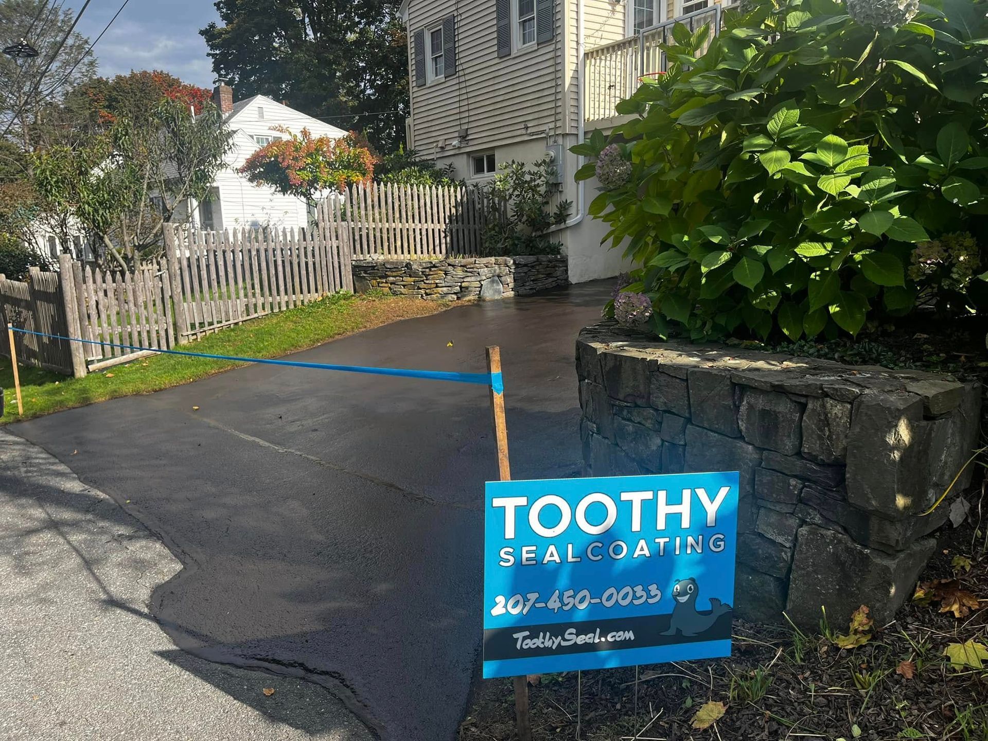 Blue Toothy Seal Coating sign on a driveway beside a stone wall and white fence