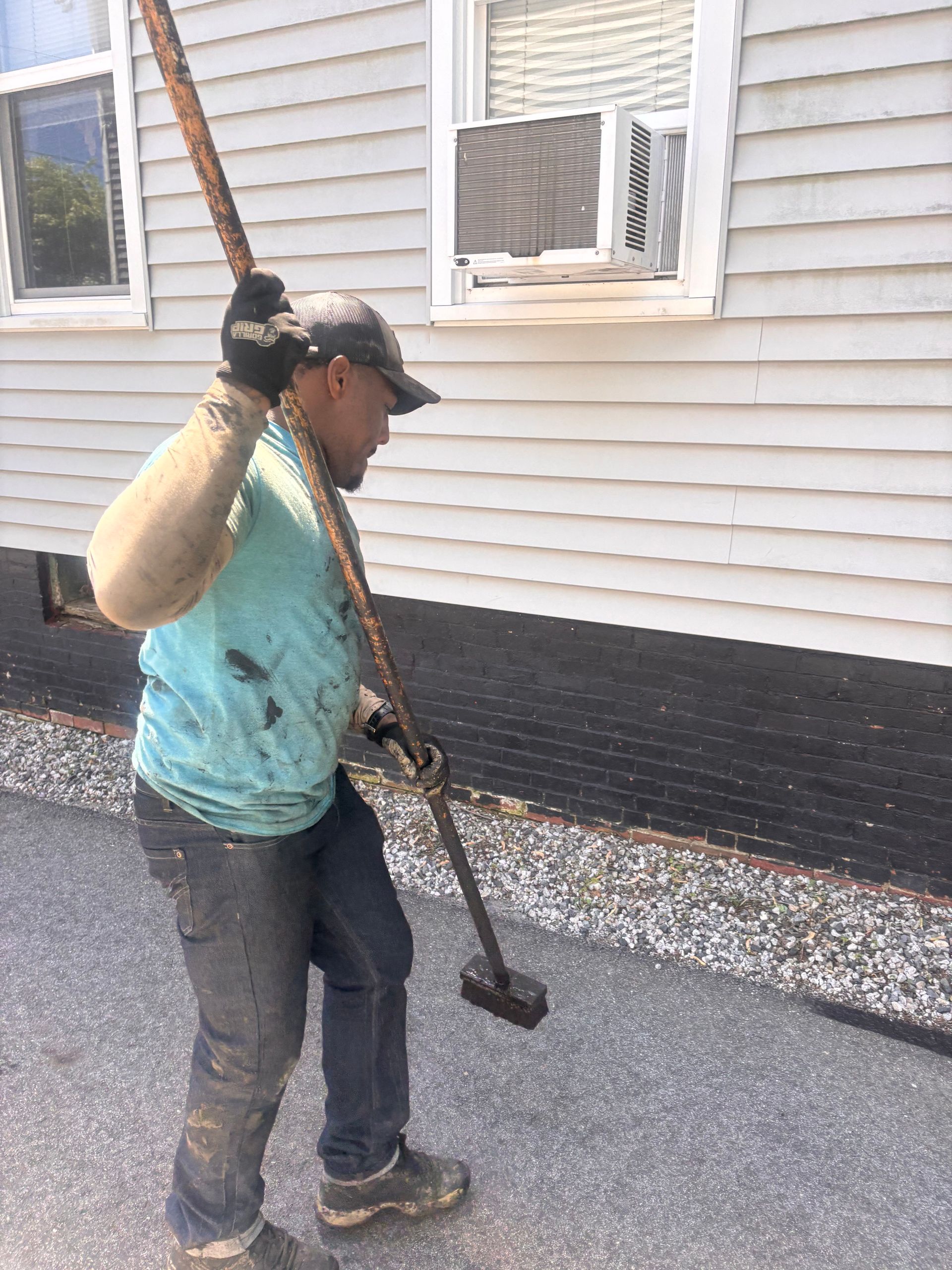 Worker carrying a long-handled tool beside a house with white siding and a gravel border