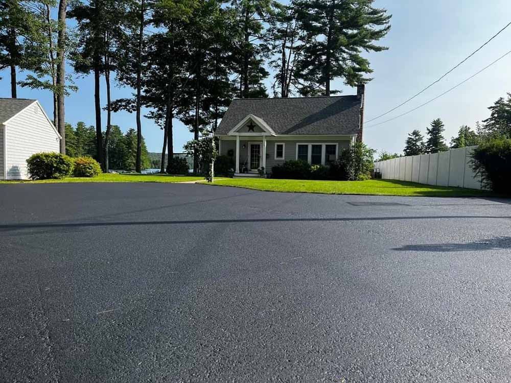 Paved driveway leading to a small white house with a porch, surrounded by trees and a white fence.