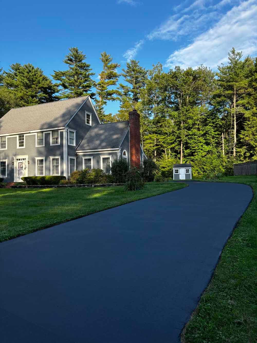 Gray house beside a long paved driveway, surrounded by green trees under a blue sky.