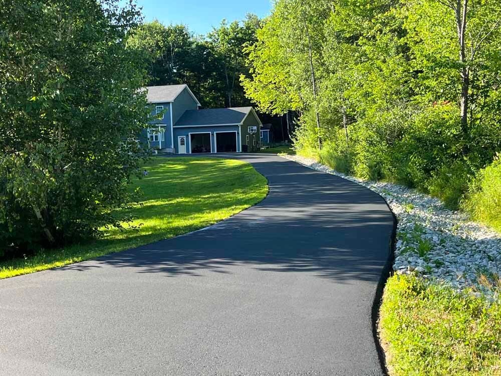 Curved driveway leading to a house, bordered by green trees and grass in bright sunlight.
