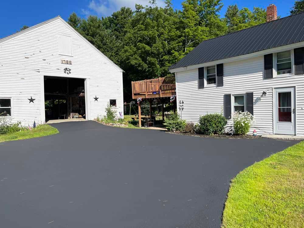 White barn and house with dark driveway and green trees under a sunny sky