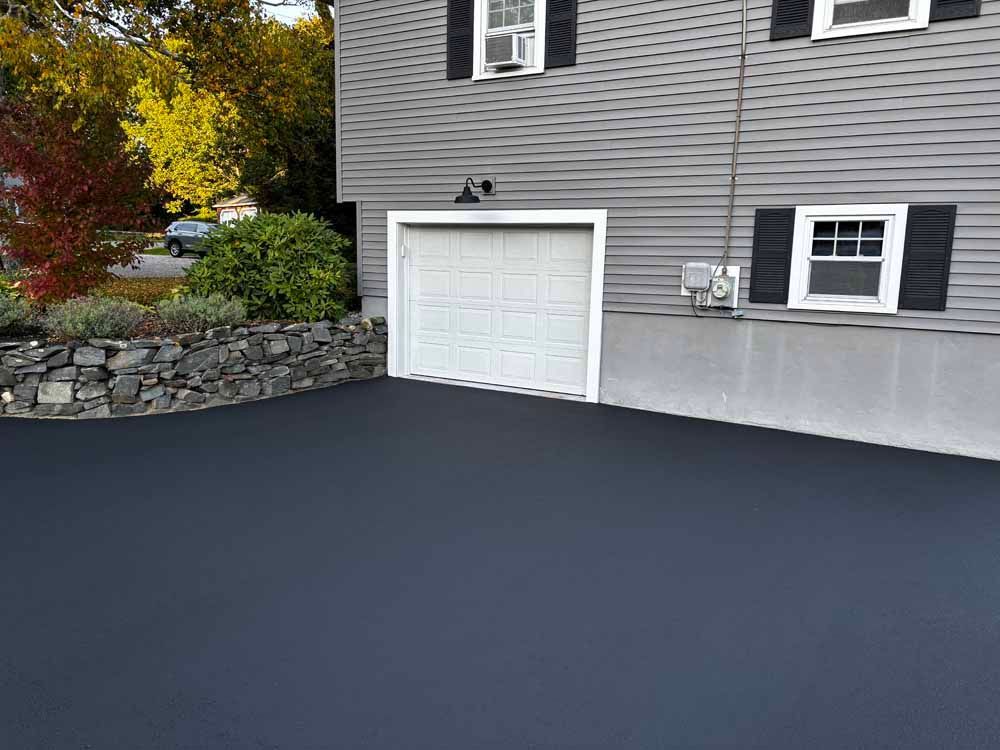 Fresh black asphalt driveway beside a gray house with a white garage door and stone retaining wall