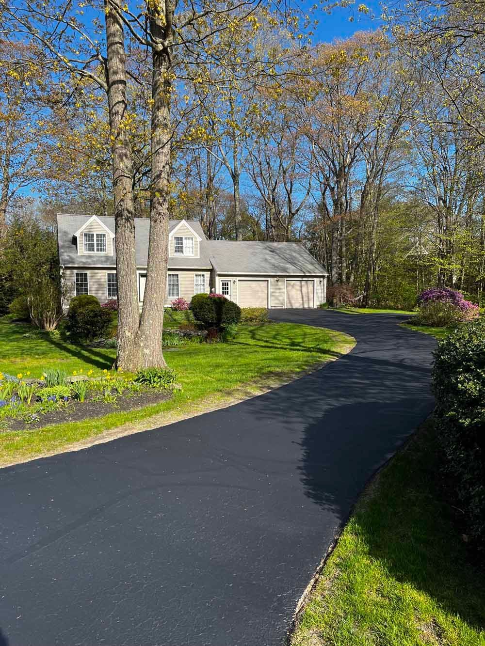 Stone house with a black driveway curving through a sunny, tree-lined front yard