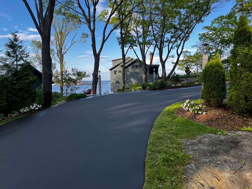 Curved paved driveway beside a lake, with trees and a house in the background.