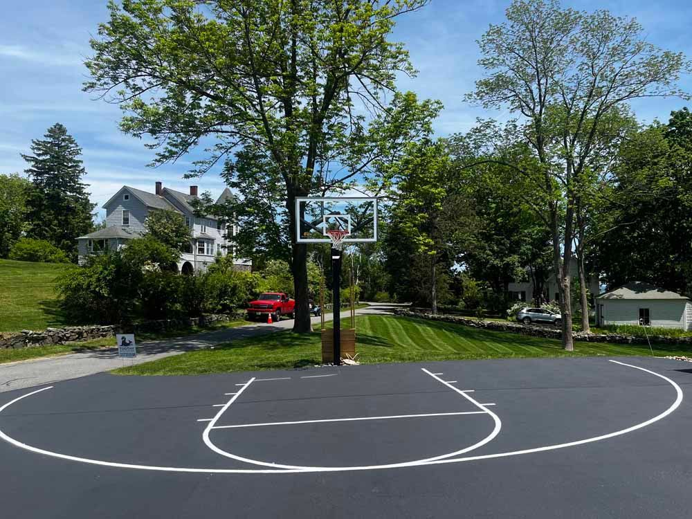 Outdoor basketball court with a hoop, surrounded by trees and a house in the background.