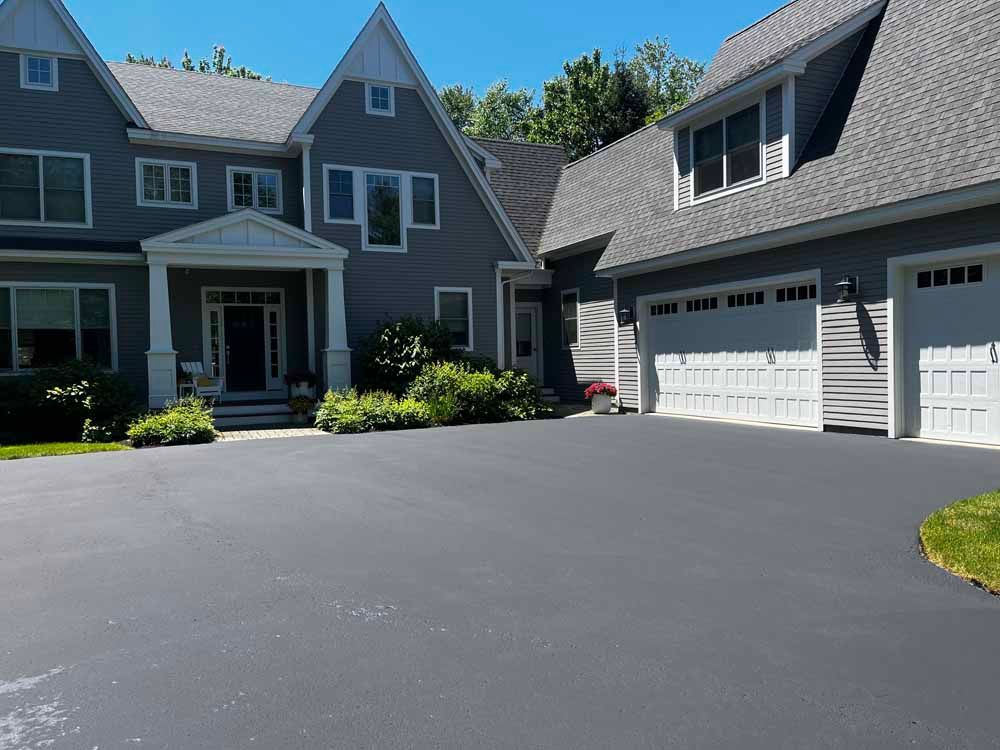 Suburban house with gray siding and attached garage, viewed from a wide paved driveway on a sunny day
