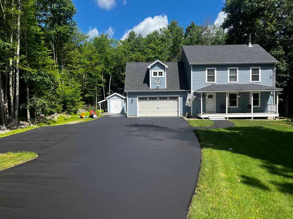 Gray two-story house with a dark roof, large driveway, and green lawn beside a wooded area