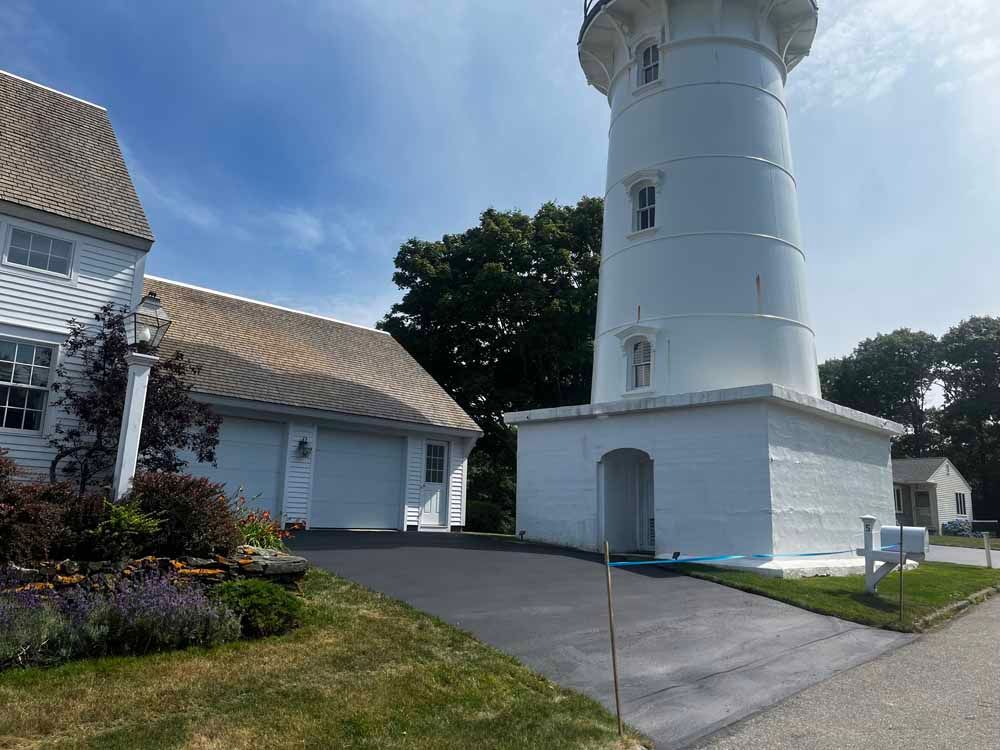 White lighthouse beside a white house with a paved driveway and manicured lawn on a sunny day