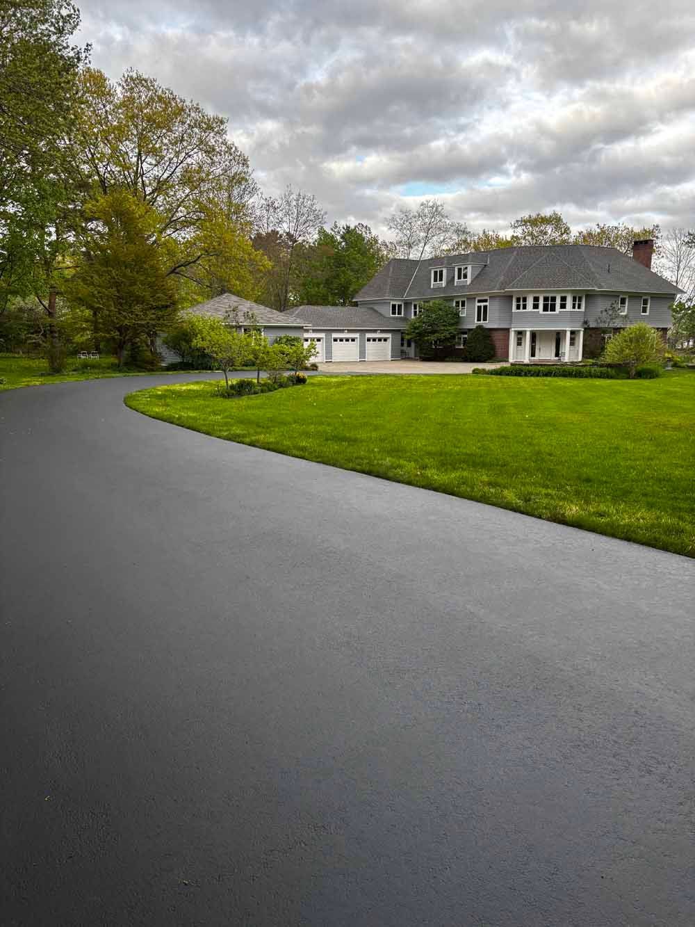 Large gray house with white garage beside a curved driveway and green lawn under cloudy skies