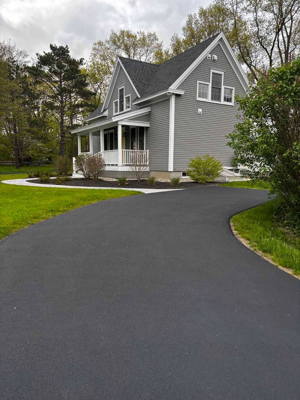Gray house with white trim beside a curved black driveway and green lawn