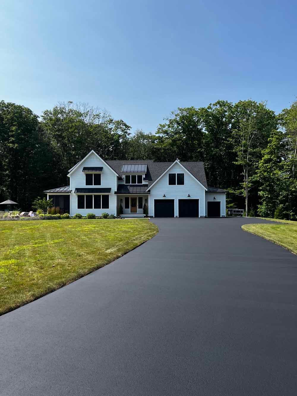 White house with two garages at the end of a long black driveway, surrounded by lawn and trees.