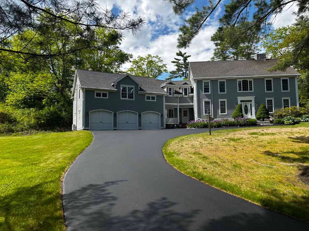 Large gray house with a two-car garage, curved black driveway, and green lawn under trees.