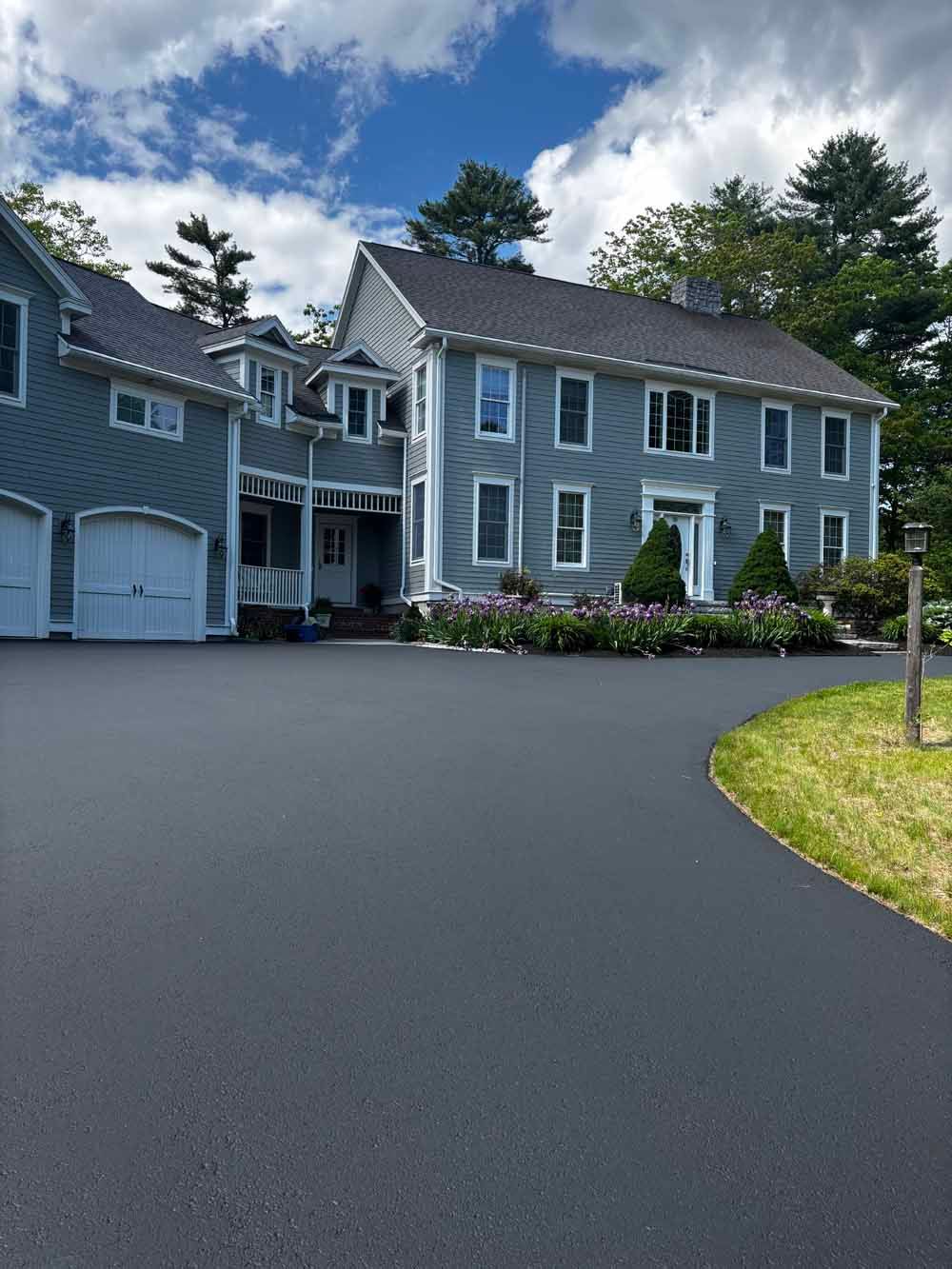 Gray house with white trim, garage, and flower beds beside a wide paved driveway under a cloudy sky