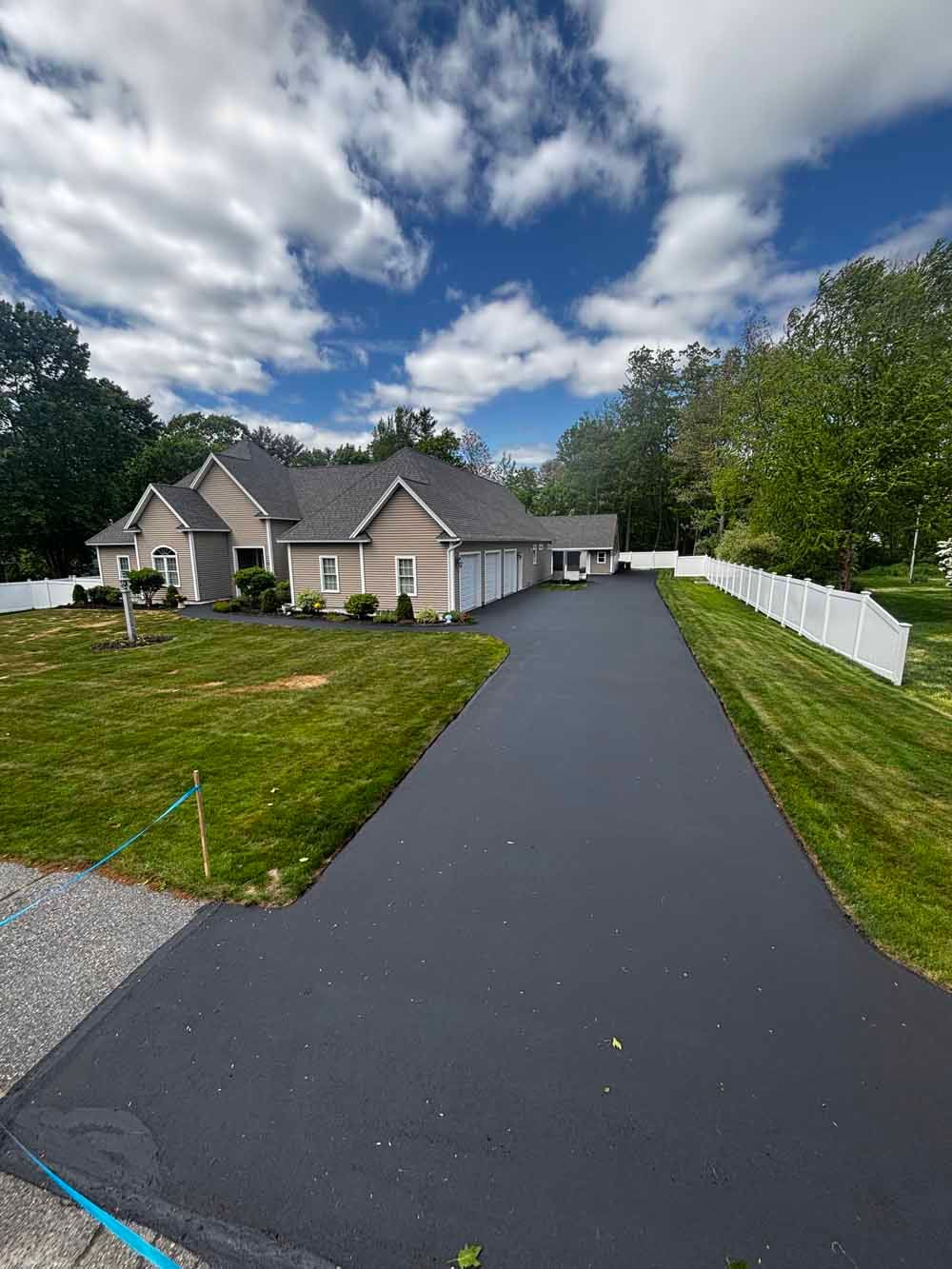 New asphalt driveway leading to a gray house with white fence and cloudy blue sky