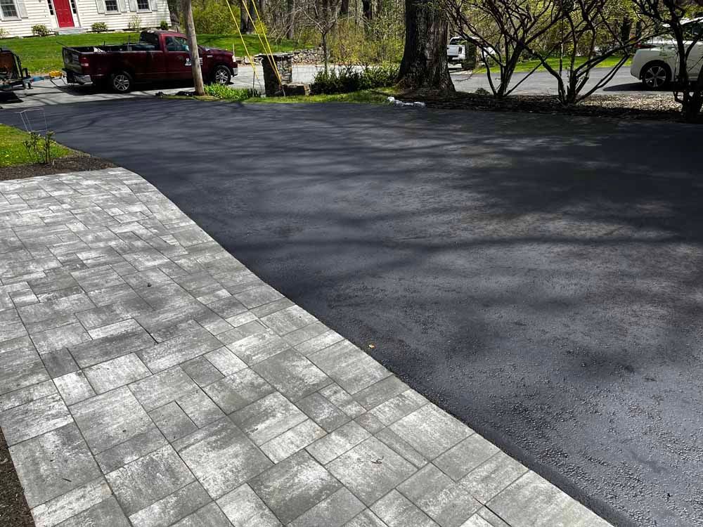 Freshly paved black driveway beside a gray stone walkway near a house and trees