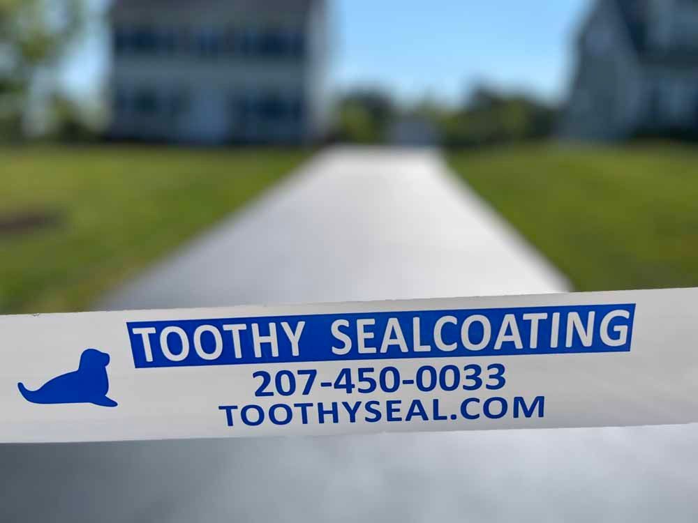Blue-and-white Toothy Sealcoating sign with phone number and website on a blurred driveway background