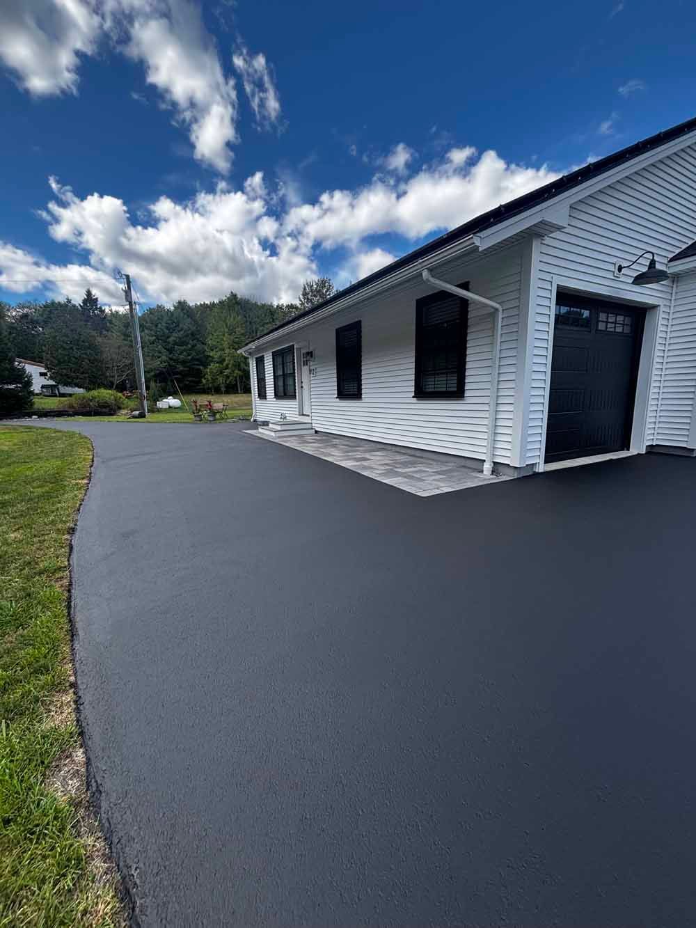 White building with black shutters beside a freshly paved driveway under a blue sky with clouds