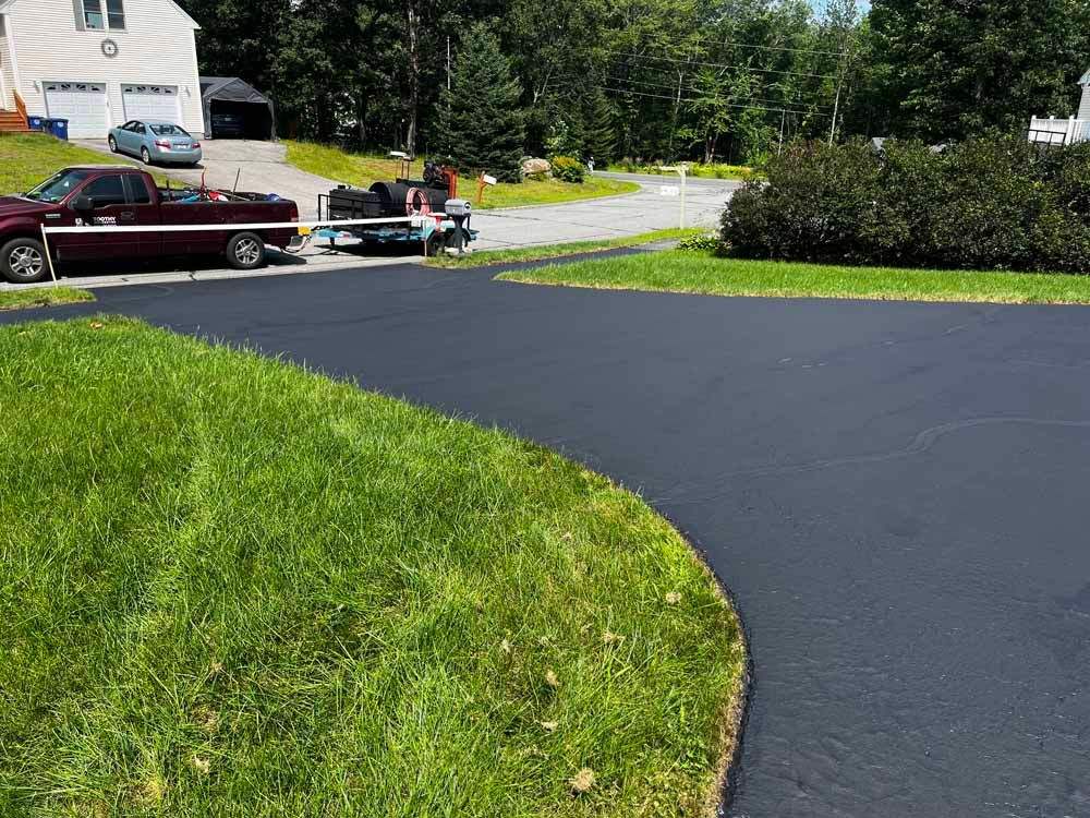 Fresh black asphalt driveway beside a red truck towing a machine, with green lawn and trees nearby