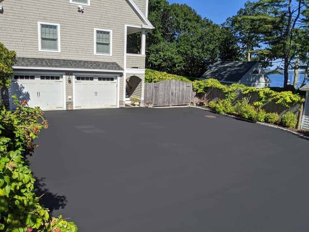 Fresh black asphalt driveway beside a gray house with white garage doors, landscaped edges, and a lake view.