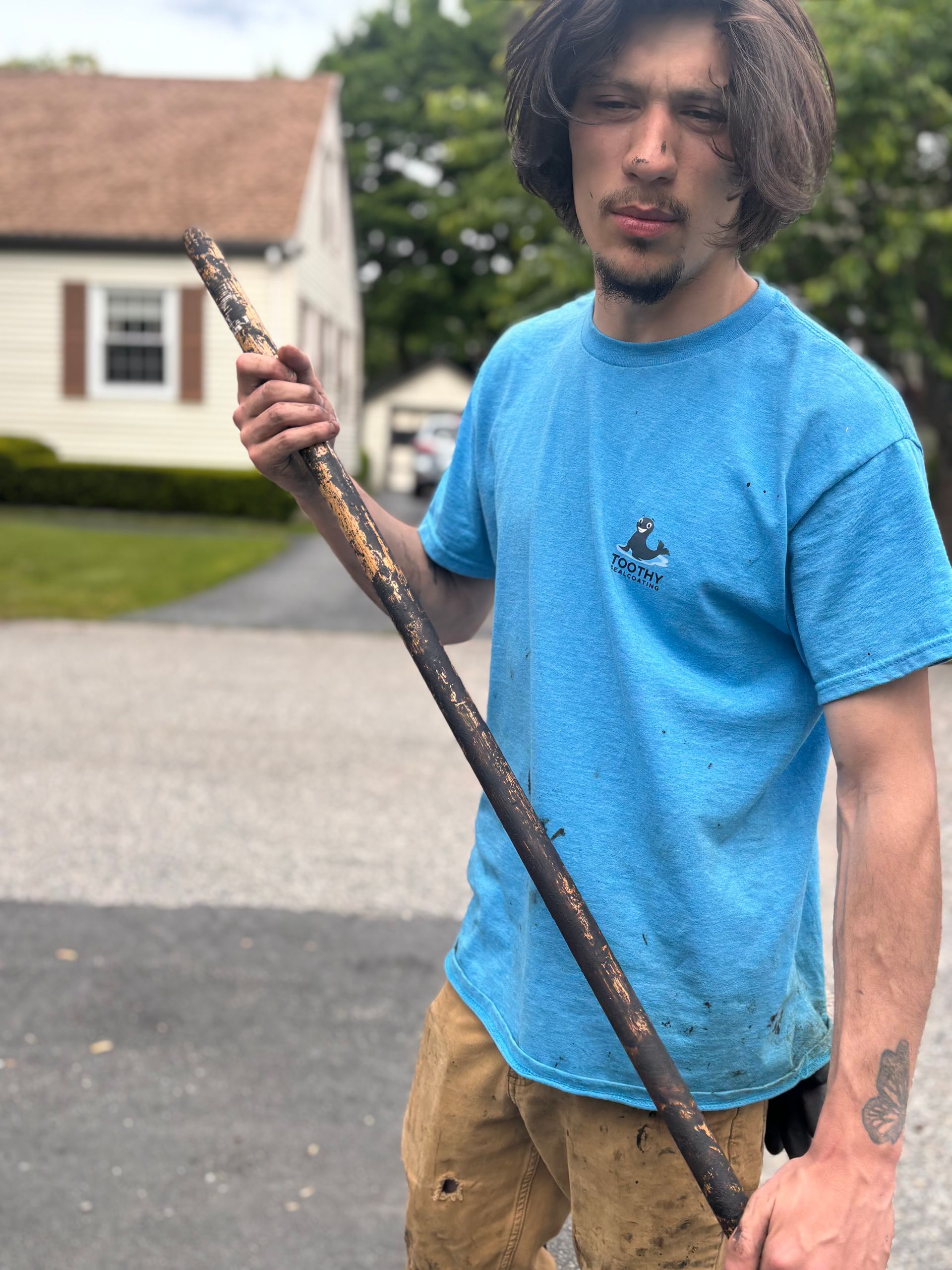 Person holding a long stick in a suburban street, wearing a bright blue shirt and tan pants.