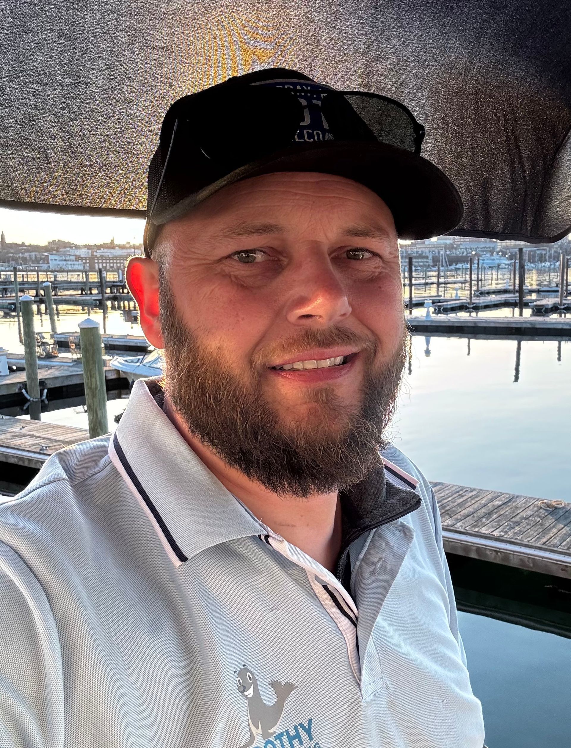 Man smiling on a dock at sunset, wearing a black cap and light gray shirt, with marina boats in the background