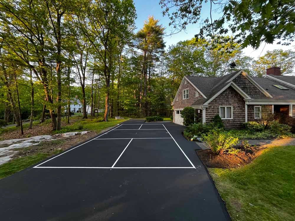 Freshly paved black tennis court beside a stone house and wooded yard