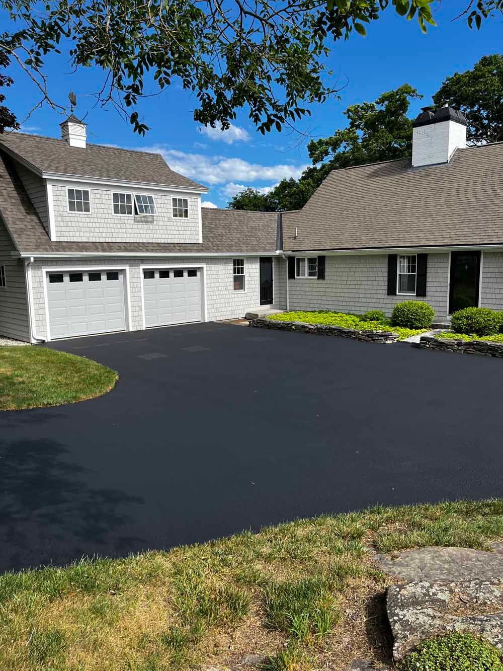 Suburban house with white siding, dark driveway, garage, and blue sky overhanging trees