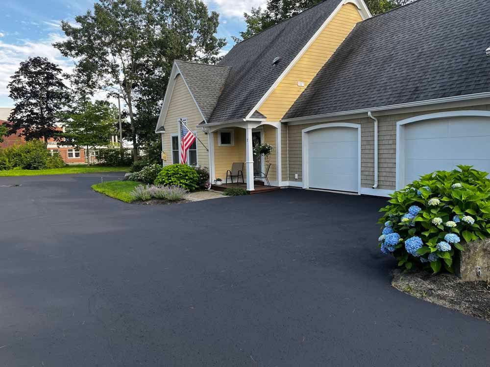 Apartment-style building with a dark roof, white garage door, and blue hydrangeas beside a paved parking lot