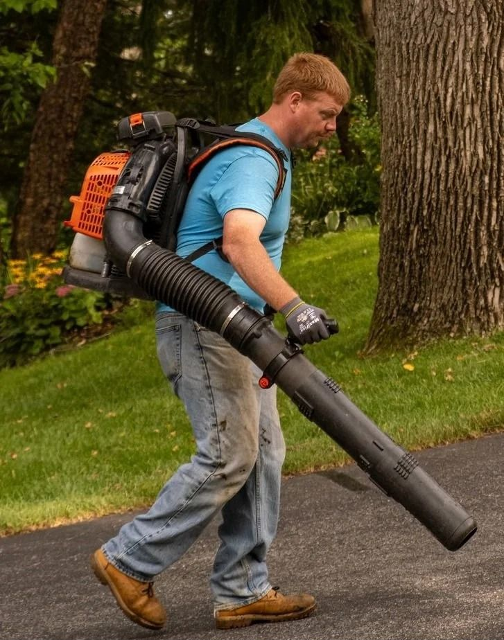 Worker using a backpack leaf blower on a paved path near grass and trees