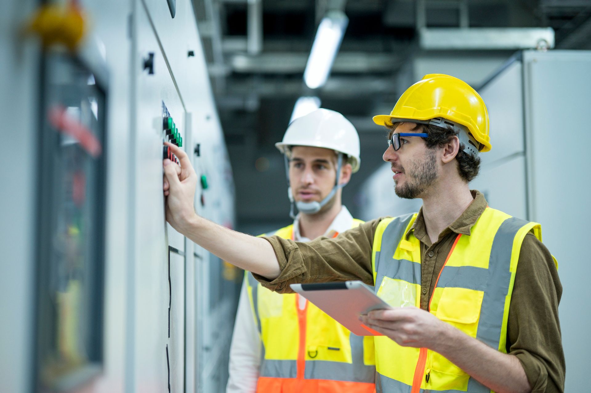Two engineers in safety vests and hard hats inspecting electrical panel in an industrial setting.