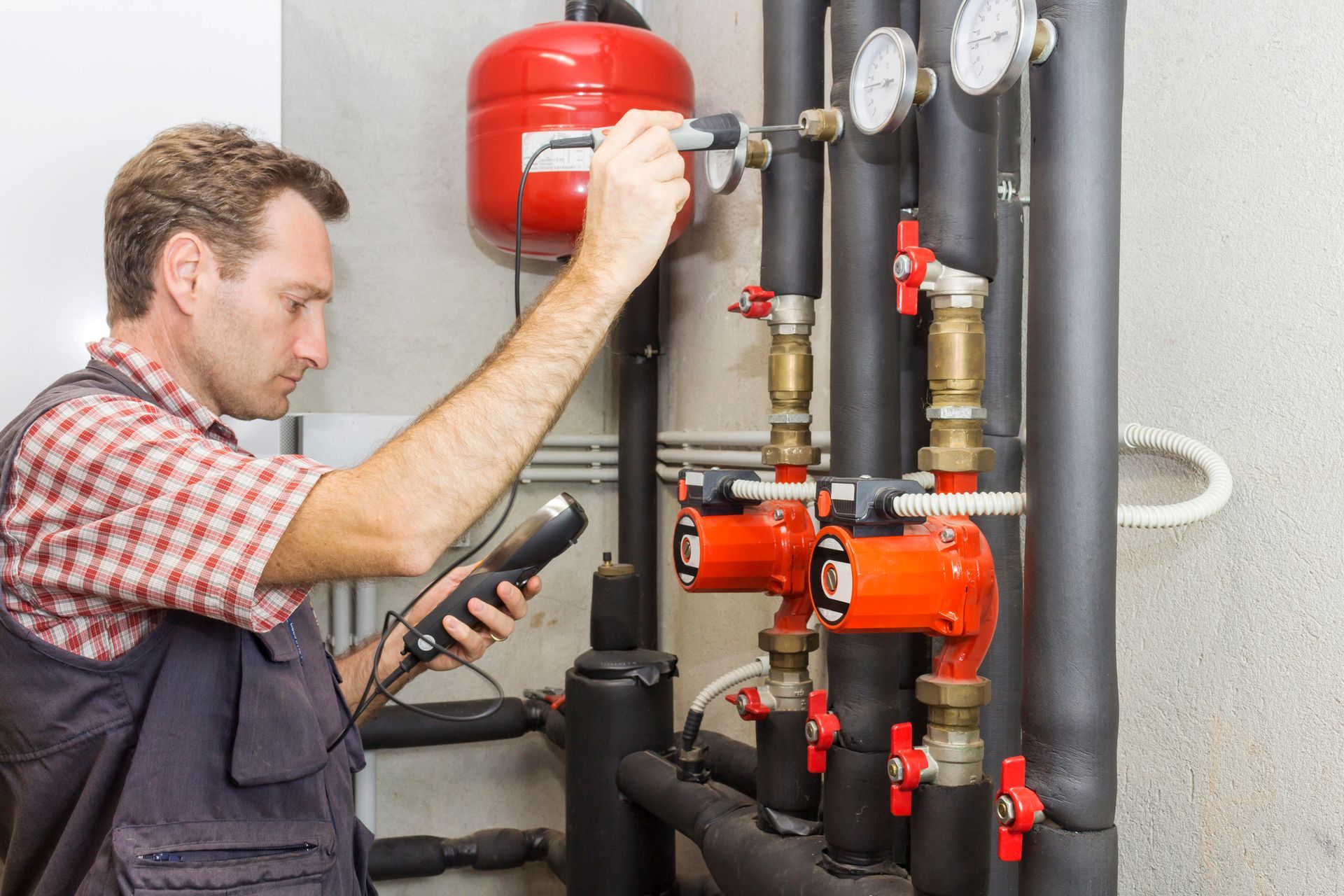 Man inspecting pipes with a device, possibly for leaks, in a mechanical room with gauges and a red tank.
