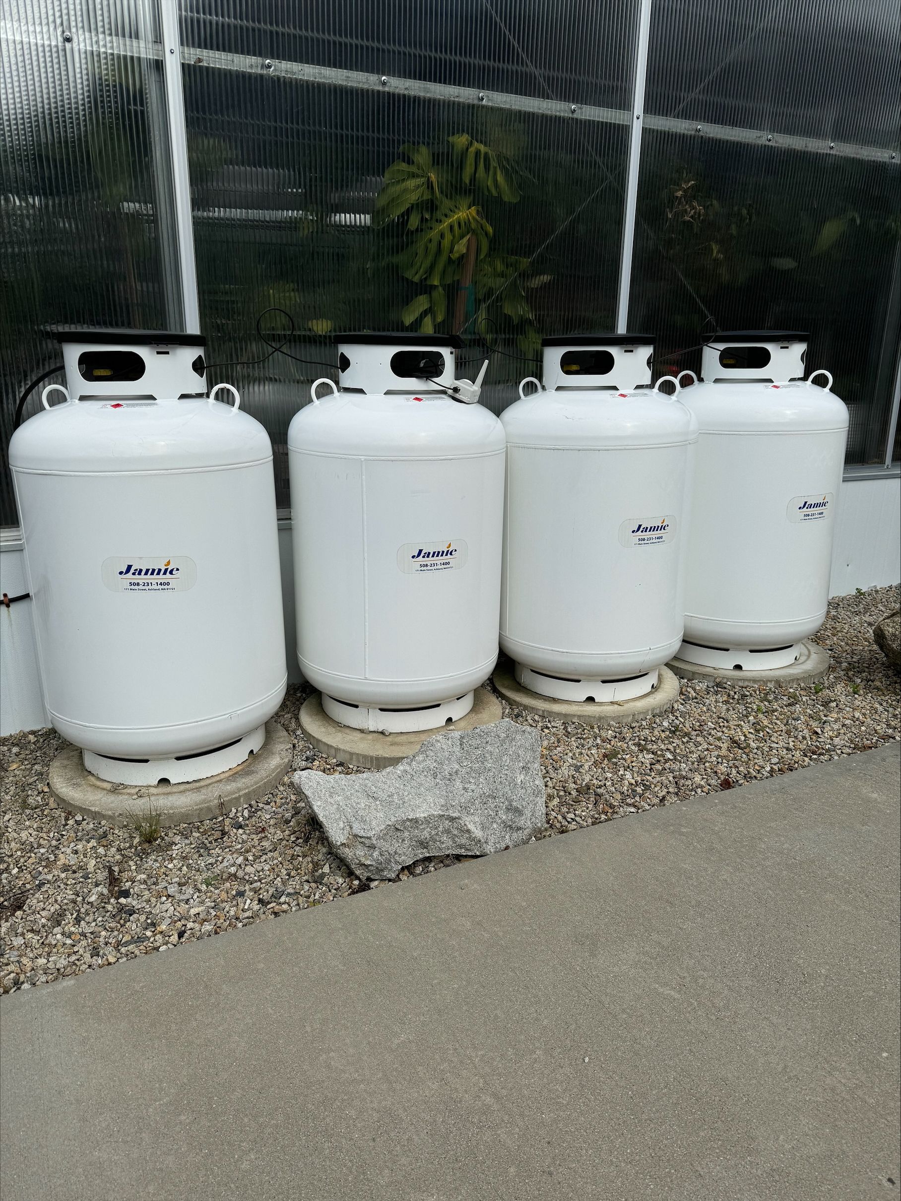 Four white propane tanks lined up beside a fence on gravel near a paved path.